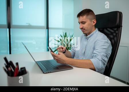 Porträt eines jungen Geschäftsmannes, der im Büro mit dem Finger auf sein Smartphone zeigt. Selbstbewusster Unternehmer, der mit dem Telefon und Laptop arbeitet Stockfoto