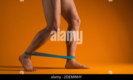 Muskulöse Männerbeine mit Gummiband für Fitness, machen Übungen, um die Beinmuskulatur zu belasten. Körperteile, gelber Hintergrund. Stockfoto