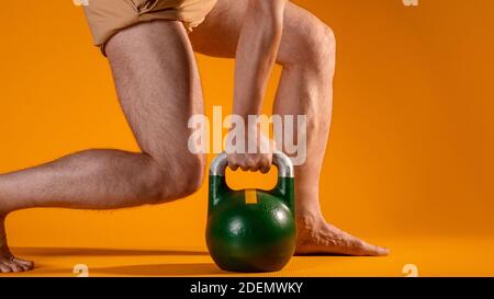 Muskulöse Männerbeine in Hocke und eine Kugelhantel in den Händen, schwingen die Beinmuskulatur, Übungen mit zusätzlichem Gewicht. Beine Nahaufnahme, gelber Hintergrund. Stockfoto