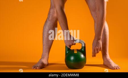 Muskulöse Männerbeine in Hocke und eine Kugelhantel in den Händen, schwingen die Beinmuskulatur, Übungen mit zusätzlichem Gewicht. Beine Nahaufnahme, gelber Hintergrund. Stockfoto