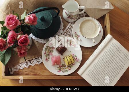 CORK, IRLAND - Nov 18, 2020: Kaffee und Mini-Kuchen auf einem Vintage-Tee-Set mit rosa Rosen, Eisen-Teekanne und ein Buch zum Lesen serviert. Stockfoto