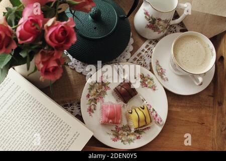 CORK, IRLAND - Nov 18, 2020: Kaffee und Mini-Kuchen auf einem Vintage-Tee-Set mit rosa Rosen, Eisen-Teekanne und ein Buch zum Lesen serviert. Stockfoto
