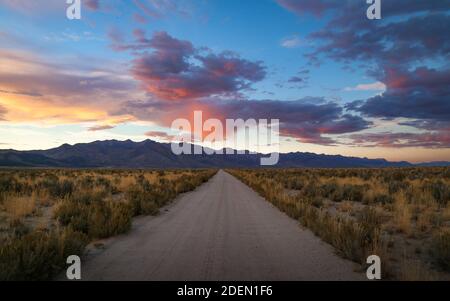 Ein dramatischer, farbenfroher Wüstenuntergang mit einer leeren Schotterstraße, die zu den fernen Bergen führt. Stockfoto
