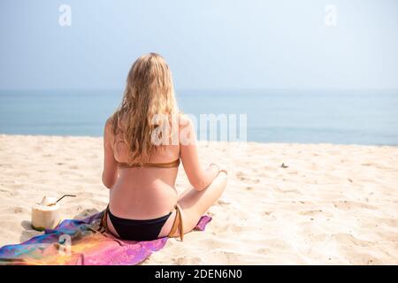 Kaukasische Frau im Bikini sitzt auf dem Sand am Strand in der Lotusposition. In der Nähe gibt es eine Kokosnuss zum Trinken. Reisen und Tourismus. Yoga-Klasse Stockfoto