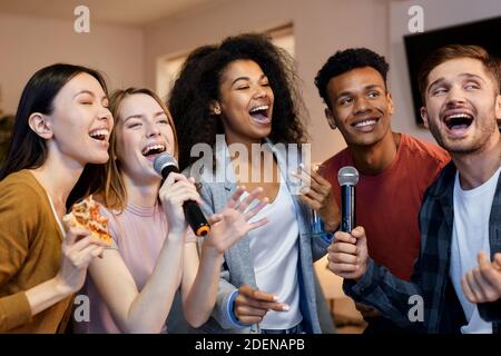 Singen Sie es. Enthusiastische Gruppe von Freunden, die mit Mikrofon singen, während sie Karaoke zu Hause spielen und in der modernen Wohnung stehen Stockfoto