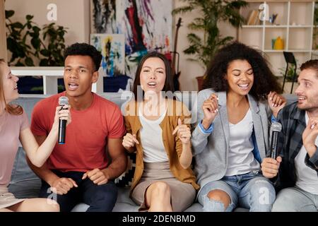 Musik bringt Freude. Enthusiastische multikulturelle Freunde singen mit Mikrofon, während sie Karaoke zu Hause spielen, sitzen auf der Couch in der modernen Stockfoto