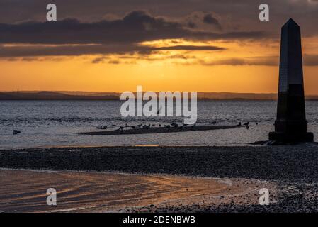 Southend on Sea, Essex, Großbritannien. Dezember 2020. Der erste Tag im Dezember ist zu Ende, als er begann; hell, aber sehr kalt. Die Sonne unterging hinter einer Wolkenschicht hinter dem Crowstone im Stadtteil Chalkwell von Southend. Der Crowstone ist ein historischer Markerstein, auch bekannt als London Stone, in der Themse Mündung, die früher die Grenzen der Gerichtsbarkeit der City of London markiert und stammt aus dem Jahr 1837. Steine standen auf dem Gelände aus den 1200er Jahren. Seevögel Stockfoto