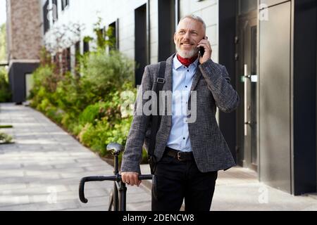 Stilvoller, reifer Geschäftsmann, der lächelt, während er am Telefon telefoniert und tagsüber mit dem Fahrrad im Freien läuft Stockfoto