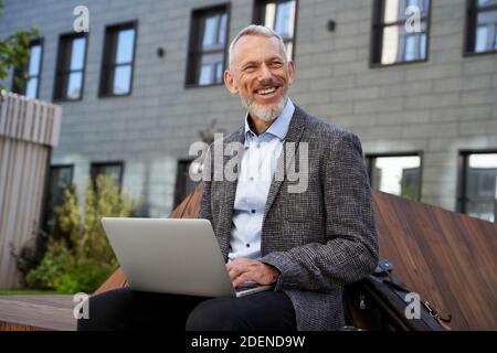 Geschäftsideen. Portrait von fröhlichen eleganten mittleren Alters Geschäftsmann lächelnd, während der Arbeit auf seinem Laptop, sitzt auf der Bank in der Nähe von modern Stockfoto