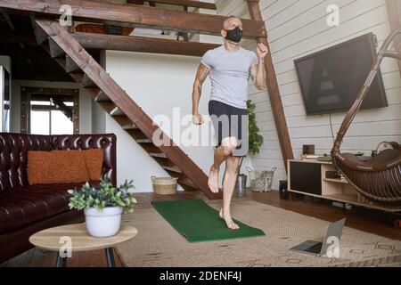 Sportlicher Mann mittleren Alters trägt Gesichtsschutzmaske Ausübung in Selbstisolierung beim Online-Workout auf dem Laptop im Wohnzimmer von ihm Stockfoto