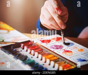 Ein Künstler hält einen Pinsel in der Hand, der Aquarellfarbe auf einer Palette mischt, und daneben stehen verschiedene Farben von Farbe und Papier, beleuchtet von Stockfoto