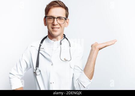 Porträt eines jungen Arztes, auf weißem Hintergrund, der die Hand auf einen leeren Blick zeigt, Auszubildender in der Werkstatt, mit Stethoskop am Hals, Stockfoto