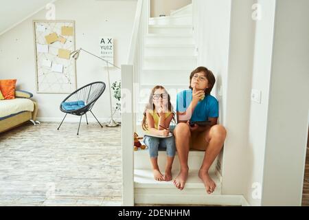 In voller Länge Aufnahme von kleinen Bruder und Schwester tragen Brillen Zu Hause auf der Treppe sitzen und dabei an etwas denken Gemeinsam Hausaufgaben machen Stockfoto