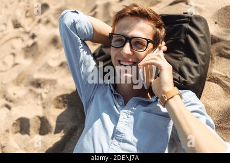 Junger Mann mit Brille, am Strand auf dem Sand liegend und telefoniert, Arbeit im Urlaub, Stockfoto