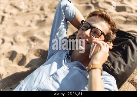 Junger Mann mit Brille, am Strand auf dem Sand liegend und telefoniert, Arbeit im Urlaub, Stockfoto