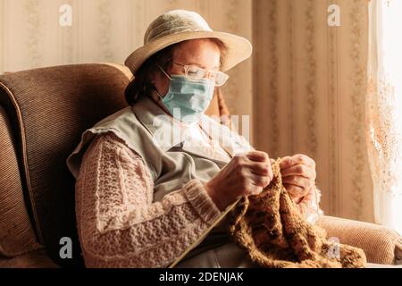 Ältere Frau mit einer medizinischen Maske Strickpullover für sie Enkel am Fenster Stockfoto