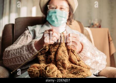 Ältere Frau mit einer medizinischen Maske Strickpullover für sie Enkel am Fenster Stockfoto