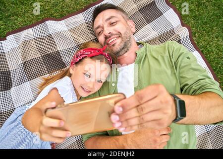 Nettes kleines Mädchen verbringt Zeit mit ihrem Vater, Selfie mit dem Smartphone, während sie sich auf einer Decke im schönen grünen Park auf einem warmen Stockfoto