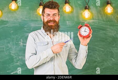 Bärtige hipster hält, schwarzes Brett für den Hintergrund. Mann mit Bart und Schnurrbart auf lächelnde Gesicht steht im Klassenzimmer. Lehrer in Brillen hält Wecker und Stift. Disziplin Konzept. Stockfoto