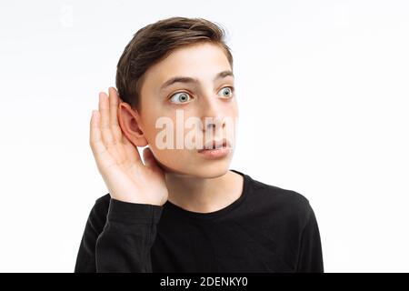 Porträt eines schönen Teenagers, neugieriger Kerl hört, macht eine Handbewegung zum Ohr, in einem schwarzen T-Shirt, auf weißem Hintergrund, Stockfoto