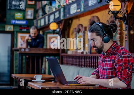 Junger bärtiger Freiberufler oder Blogger, sitzt in einer Kneipe, arbeitet an einem Laptop-Computer, trägt Kopfhörer auf dem Kopf. Ein anderer Kunde aus der Ferne. Stockfoto