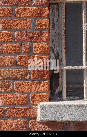 Ein vertikales Nahaufnahme-Bild eines alten, weißen, vergitterten Fensters, mit Morgenschatten, in einer orangefarbenen Ziegelwand. Stockfoto