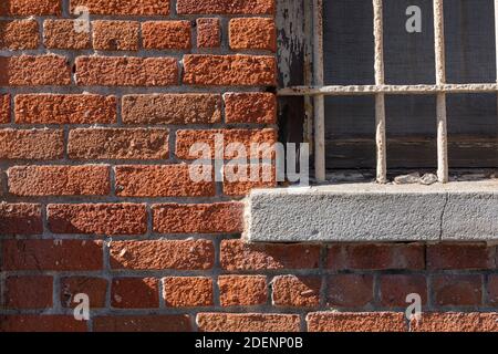 Ein horizontales Nahaufnahme-Bild eines alten, weißen, vergitterten Fensters, mit Morgenschatten, in einer orangefarbenen Ziegelwand. Stockfoto