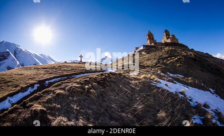 Wiesenweg zur Dreifaltigkeitskirche Gergeti mit schneebedeckten Bergen Und strahlende Sonne im Hintergrund Stockfoto