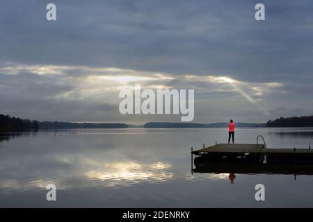 Die Sonnenstrahlen ziehen durch die Wolken über dem See, eine weibliche Silhouette auf einem hölzernen Pier, Lake Tuusula, Finnland Stockfoto