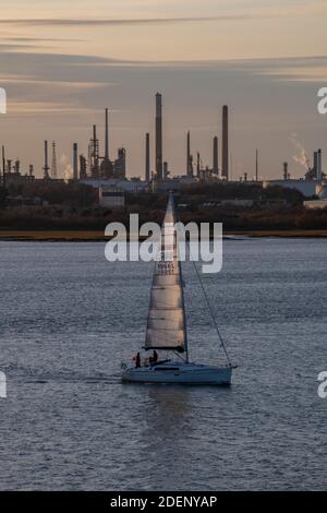 Eine Segelyacht bei Sonnenuntergang am solent vorbei an der industriellen Kulisse der fawley Raffinerie und des Kraftwerks. Stockfoto