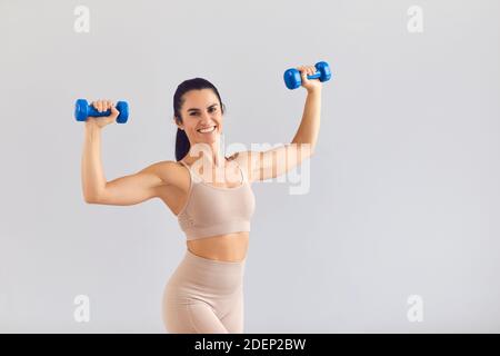 Positive sportliche Frau in beige activewear lächelnd und posiert mit Kurzhanteln im Studio Stockfoto