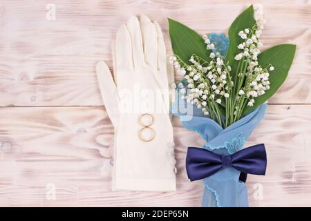 Blumenstrauß Maiglöckchen aus dem Tal in türkisfarbener Serviette, weiße Hochzeitshandschuhe und blaue Fliege auf hellem rustikalem Tisch.Hochzeitsgrusskarte Stockfoto