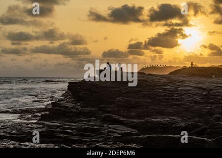 Sonnenuntergang über Accra Strand mit Felsen und zwei Personen sitzen Und genießen Sie das Meer mit hügeligen Boden, wo Sie sehen Die Schießstand Zeichen in der Hintergro Stockfoto