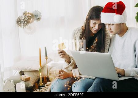 Happy stilvolle Paar kaufen Geschenke online mit Laptop und Kreditkarte, sitzen in stilvollen Boho Zimmer mit festlichen Weihnachtsdekorationen. Weihnachtsangebot A Stockfoto