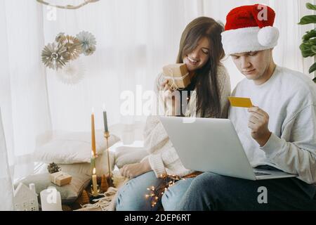 Happy stilvolle Paar online mit Laptop und Kreditkarte einkaufen, sitzen in stilvollen Boho Zimmer mit festlichen Weihnachtsdekorationen. Weihnachtsangebot und B Stockfoto