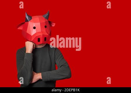 Junger Mann in roter 3d Maske einer Stierkkuh auf dem Kopf auf einem isolierten Hintergrund. Ein Mensch hält seinen Kopf, denkt. Leer für Banner, Vorlage, Platz für ein Stockfoto