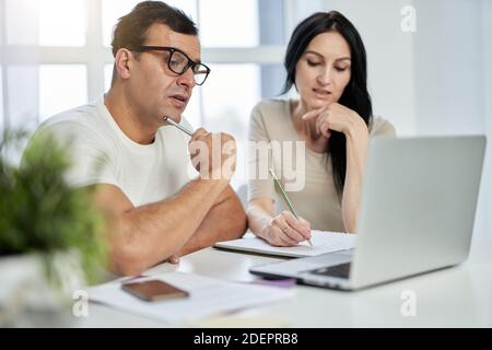 lateinisches Paar mittleren Alters, Mann und Frau, die während eines Online-Schulungskurses Notizen machen, während sie aus der Ferne lernen, mit einem Laptop zu Hause. Fernbildung für Erwachsene Stockfoto