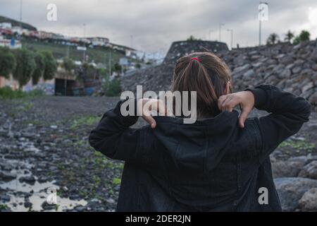 Mädchen, das an einem bewölkten Tag die Kapuze ihrer schwarzen Jacke mit beiden Händen aufsetzt. Stockfoto