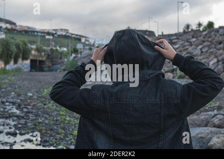 Mädchen, das an einem bewölkten Tag die Kapuze ihrer schwarzen Jacke mit beiden Händen aufsetzt. Stockfoto