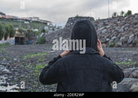 Mädchen, das an einem bewölkten Tag die Kapuze ihrer schwarzen Jacke mit beiden Händen aufsetzt. Stockfoto