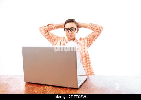 Ein junger Mann, der sich nach der Schule ausruhte, saß am Tisch und schaute sich einen Film auf einem Laptop an, warf seine Hände hinter seinen Kopf, auf einem weißen Hintergrund, hinein Stockfoto