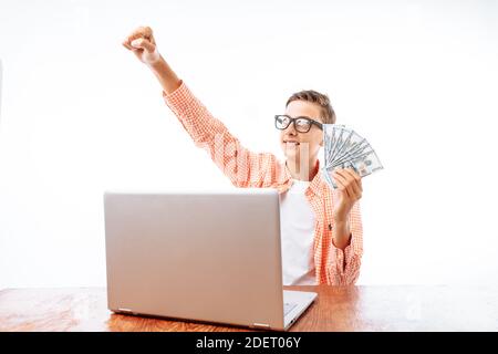 Ein junger Mann mit Geld in den Händen verdient durch das Internet, sitzt mit einem Laptop, hob seine Hand, feiert den Sieg, im Studio Stockfoto