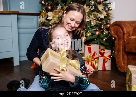 Mutter geht Überraschung für Tochter gibt Geschenk, in der Nähe Weihnachtsbaum zu Hause zu machen. Glücklich lächelndes Mädchen erhält Geschenk von der Mutter. Stockfoto