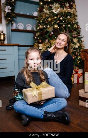 Mutter geht Überraschung für Tochter gibt Geschenk, in der Nähe Weihnachtsbaum zu Hause zu machen. Glücklich lächelndes Mädchen erhält Geschenk von der Mutter. Stockfoto