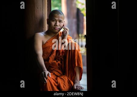 Buddhistischer Mönch in orangefarbenem Gewand ruft am Handy an, während er im Fenster sitzt, Mandalay, Myanmar Stockfoto