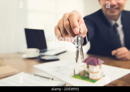 Lächelnder Geschäftsmann als Immobilienmakler Agent givng Haus Schlüssel an seinem Schreibtisch im Büro Stockfoto