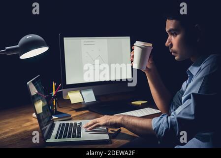 Schläfriger, müde Geschäftsmann, der spät in der Nacht Überstunden im Hotel verbracht hat Büro hält Kaffeetasse und konzentriert sich auf die Arbeit mit Laptop Computer an seinem Schreibtisch Stockfoto