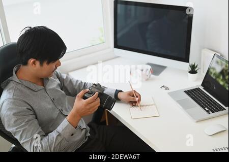 Ein junger Fotograf, der seine aufgenommenen Fotos in seiner Kamera anführt, während er im Studio an seinem Schreibtisch sitzt. Stockfoto