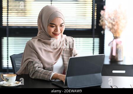 Junge arabische Unternehmerin mit einem Hijab sitzt im Büro und arbeitet online mit Tablet. Stockfoto