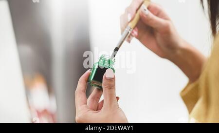 Ausschnitt einer Künstlerin, die einen Pinsel in der Hand hält und ihn in eine Farbflasche taucht. Stockfoto